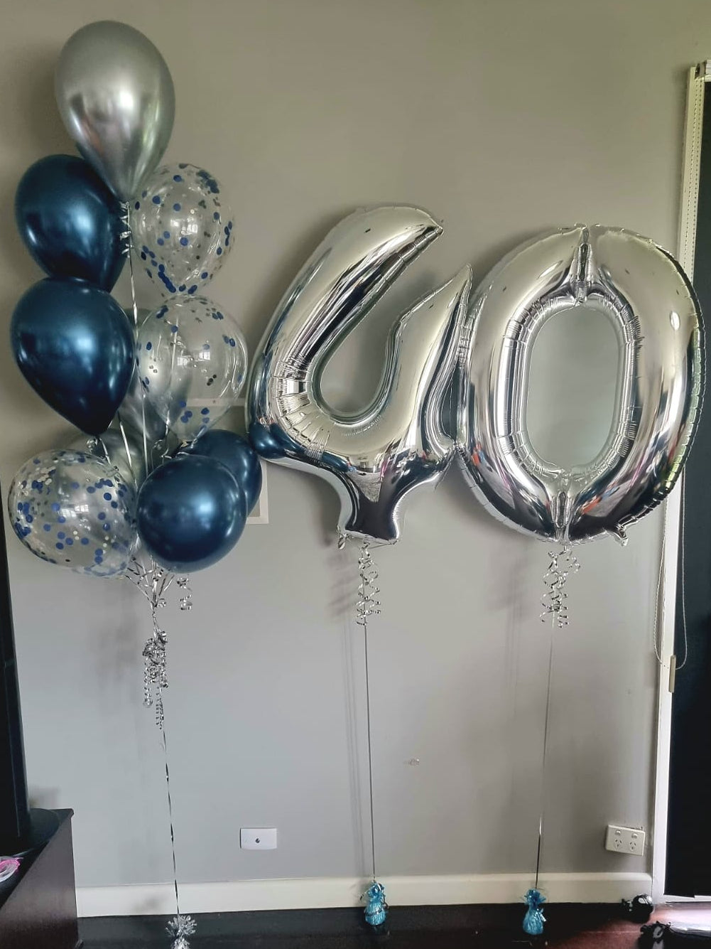 A balloon bouquet featuring large numbered balloons in silver, dark blue, and silver with smaller metallic balloons in a living room setting.