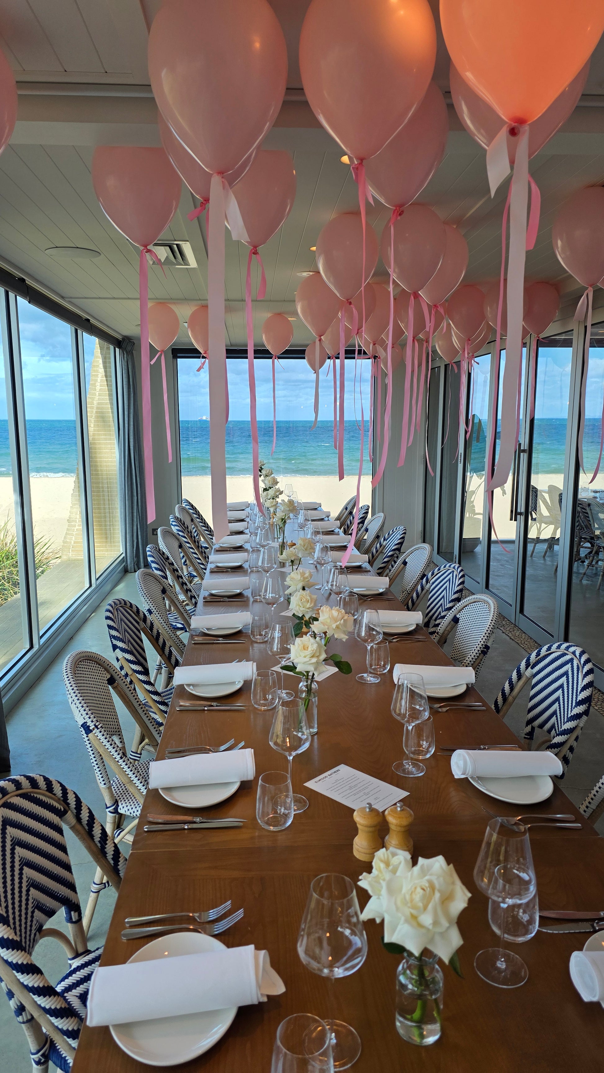 Dining table set with pink balloons and decorative items in a room with large windows.