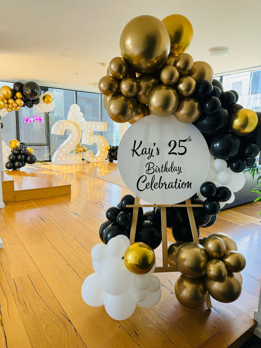 A balloon garland featuring a mix of metallic and non-metallic balloons in black, gold, and white, arranged on a gold mesh stand with a neon sign reading 'Lets Party'.