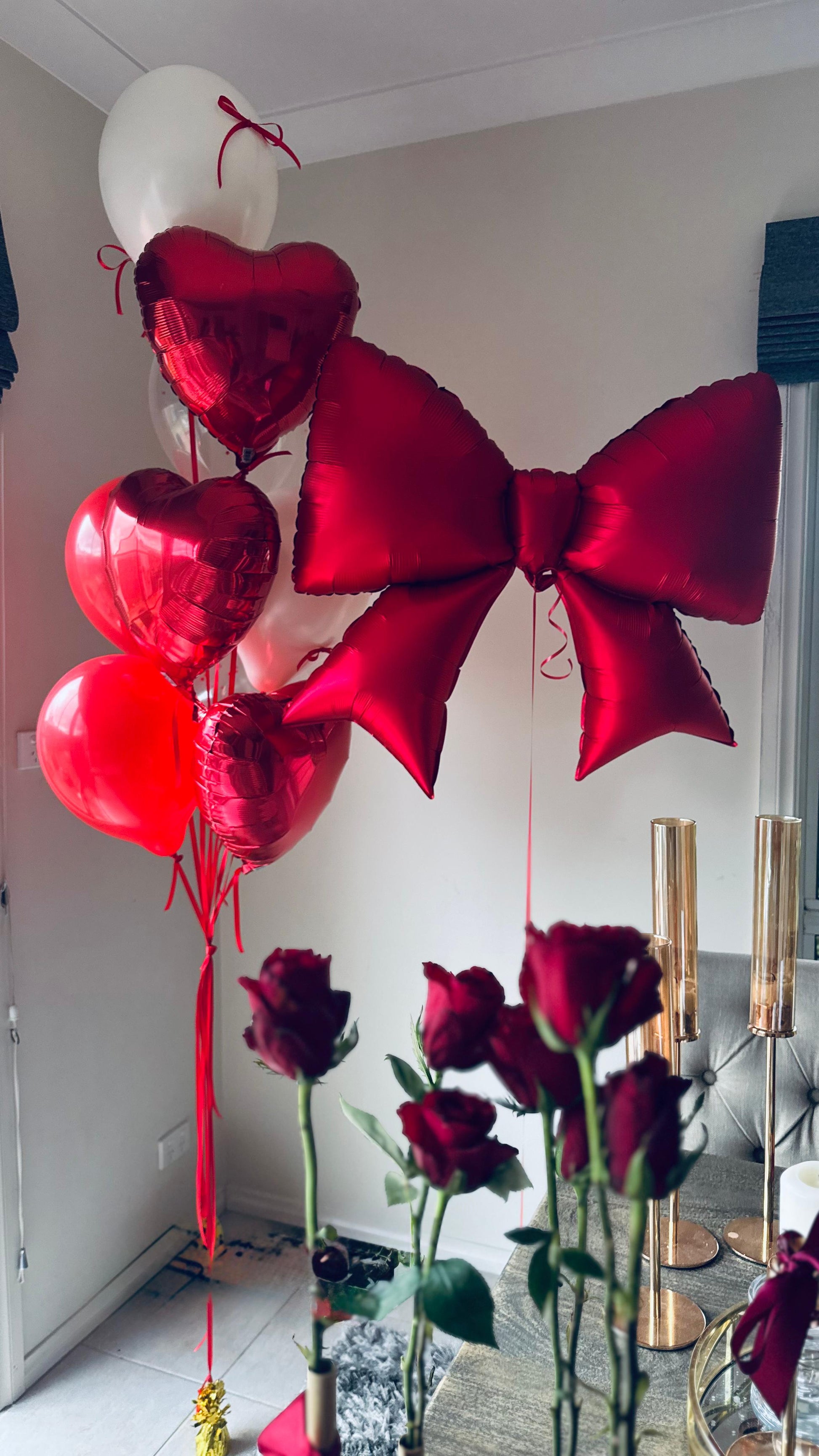 Red heart-shaped balloons and a large red bow with roses on a table.