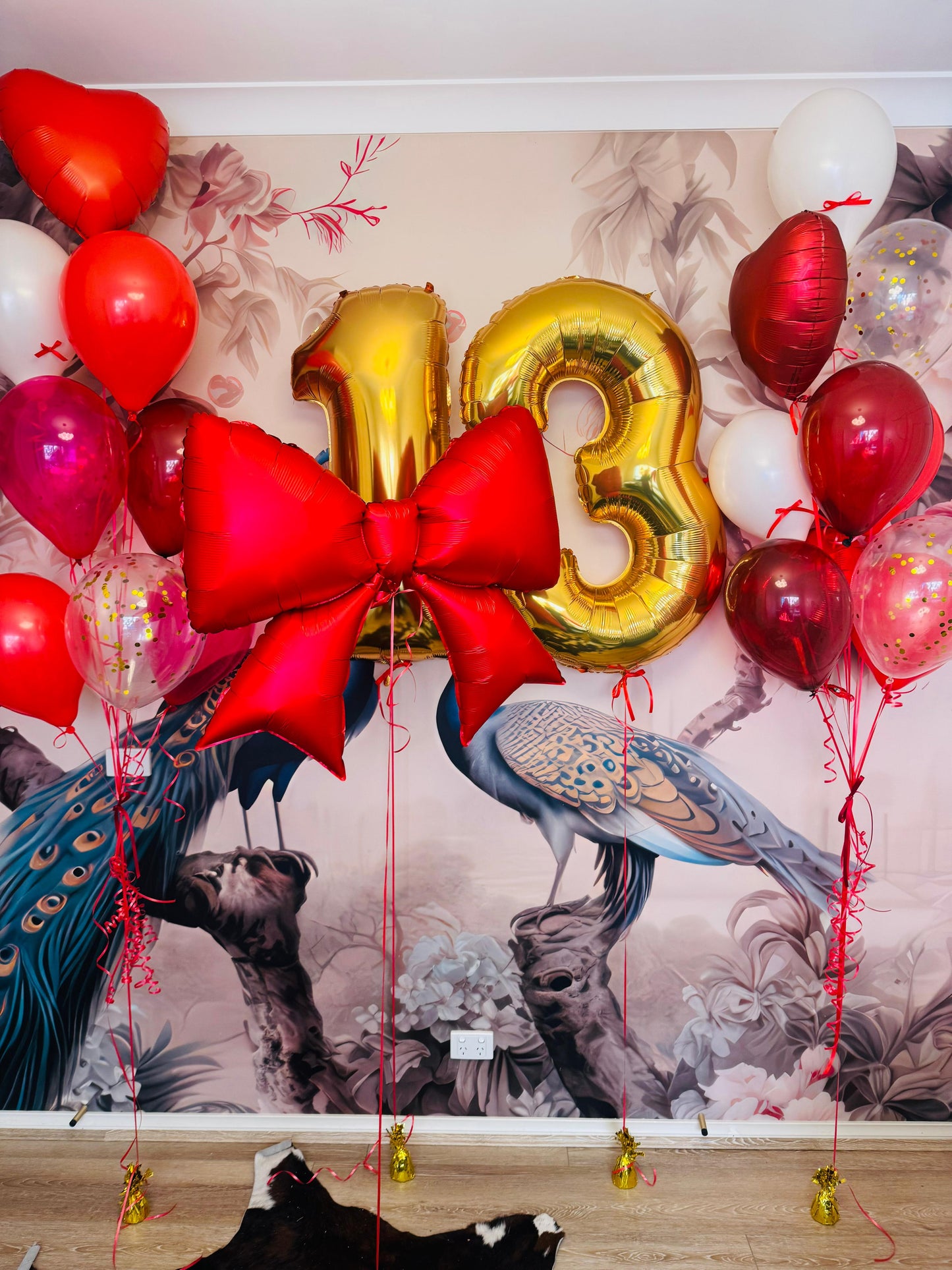 Gold number '13' balloon with a red bow and surrounding balloons against a decorative wall.