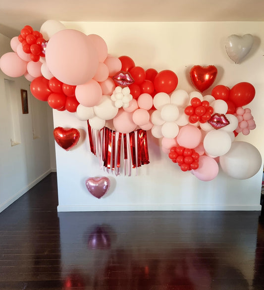 A DIY balloon garland kit with various balloons in shades of red, white, and pink, featuring hearts and tassels, displayed in a room. Shipped from Australia Globally