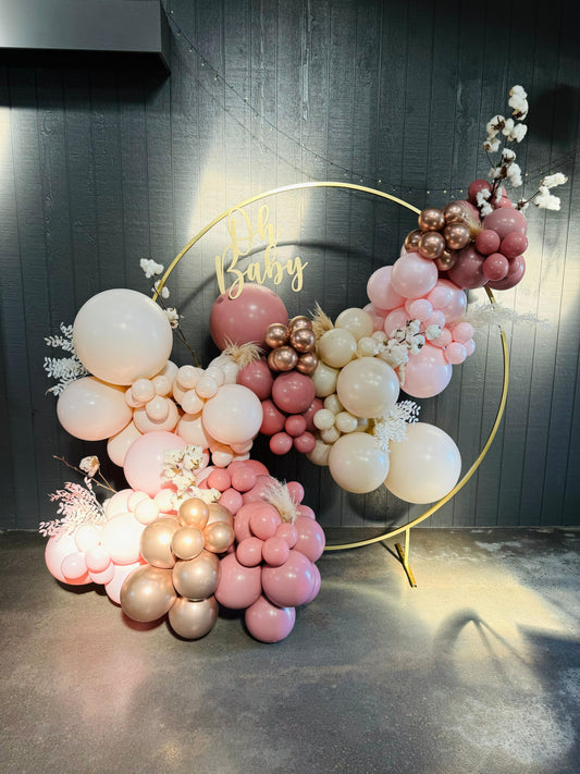 A DIY balloon garland kit with a mix of pink and peach balloons arranged in a circular frame with a golden-colored stand. The balloons are in various sizes and some are double-stuffed for a unified color appearance.