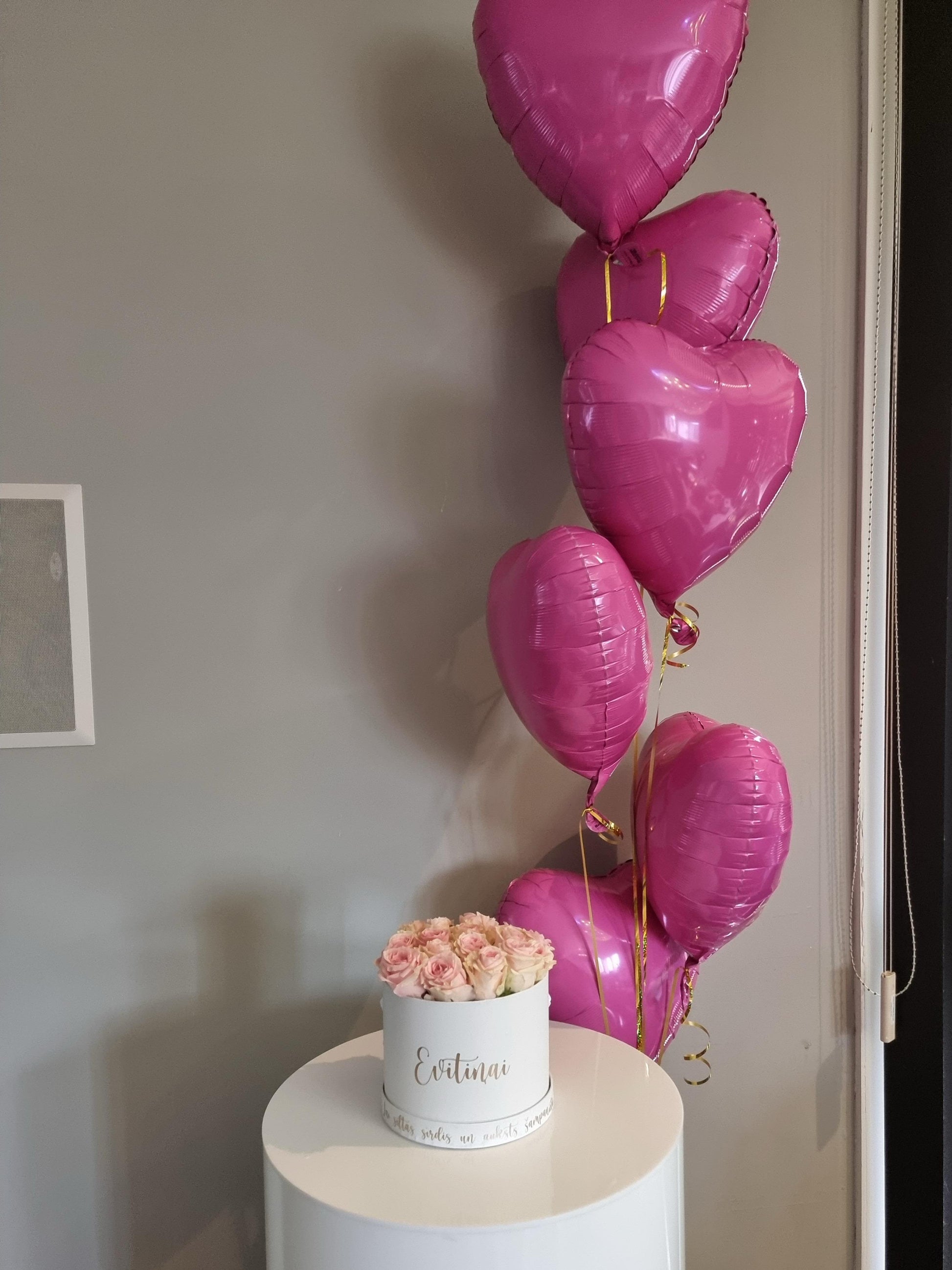 Pink roses in a box sitting on a plinth with rose coloured pink heart shaped balloons 