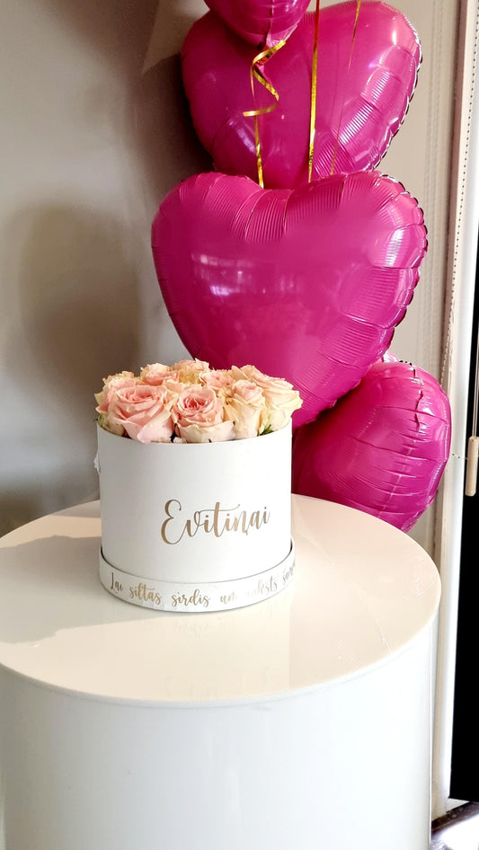 A box of pink roses with a balloon bouquet in the background, both featuring a Valentine's theme.