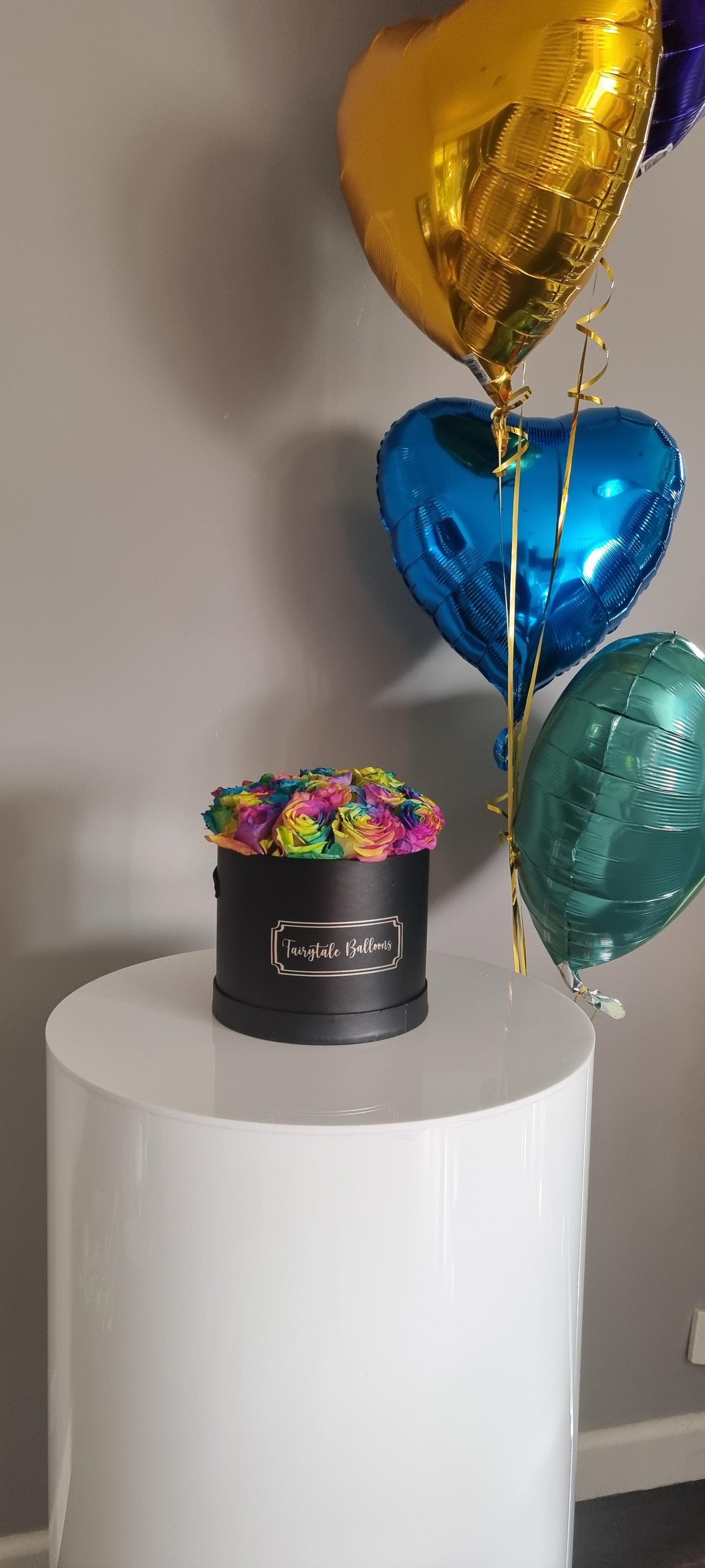 A box of pink roses with a balloon bouquet in the background, both featuring a Valentine's theme.