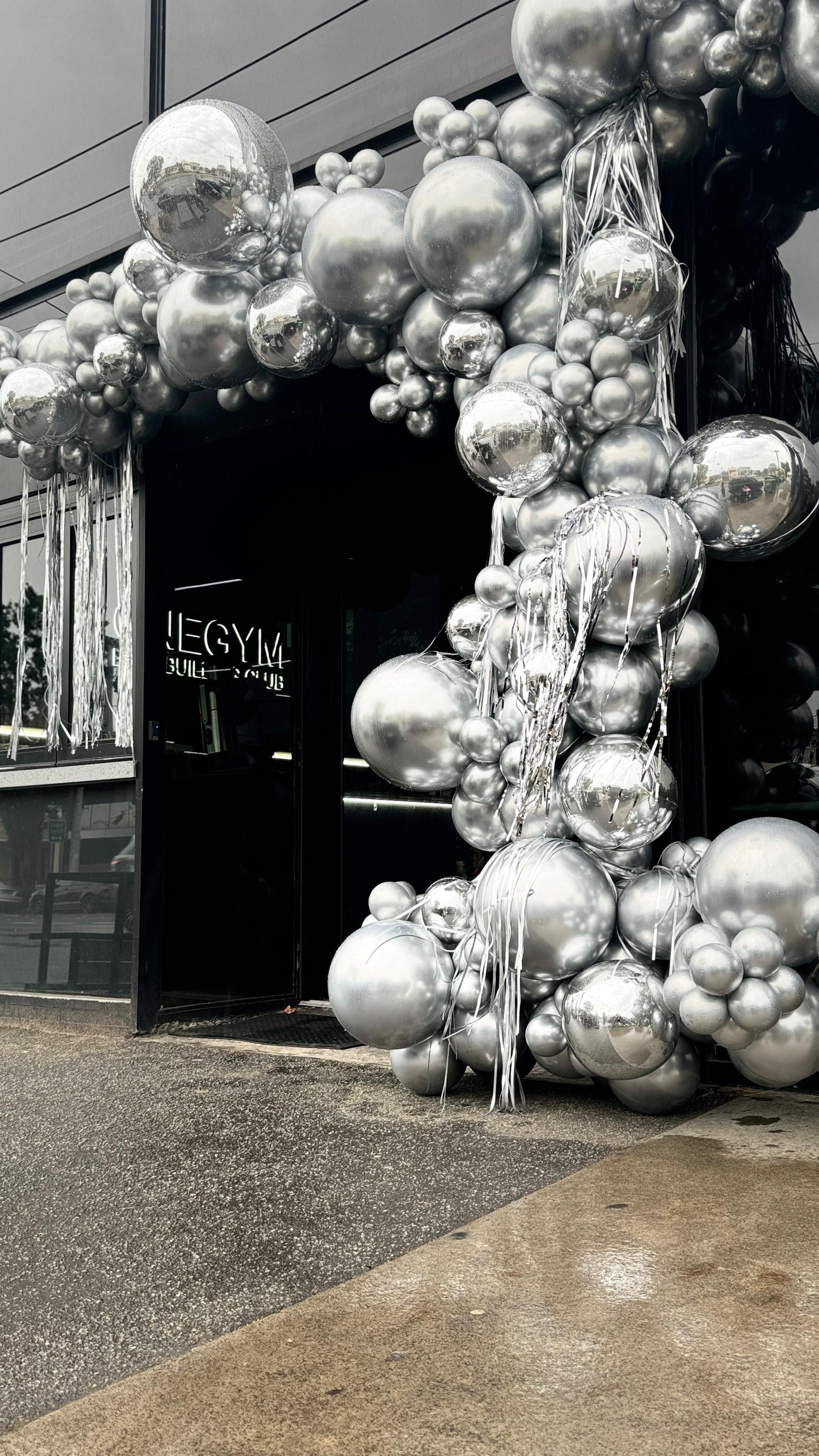 A building entrance decorated with a large, intricate balloon garland made of silver and foil balloons, with a video camera capturing the scene. Silver colour. Global Shipping