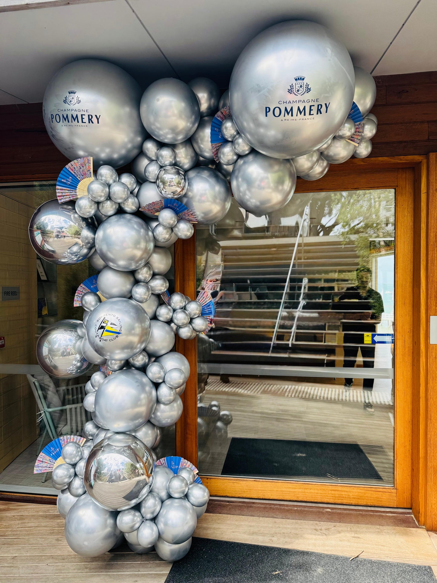 A building entrance decorated with a large, intricate balloon garland made of silver and foil balloons, with a video camera capturing the scene. Global Shipping from Australia