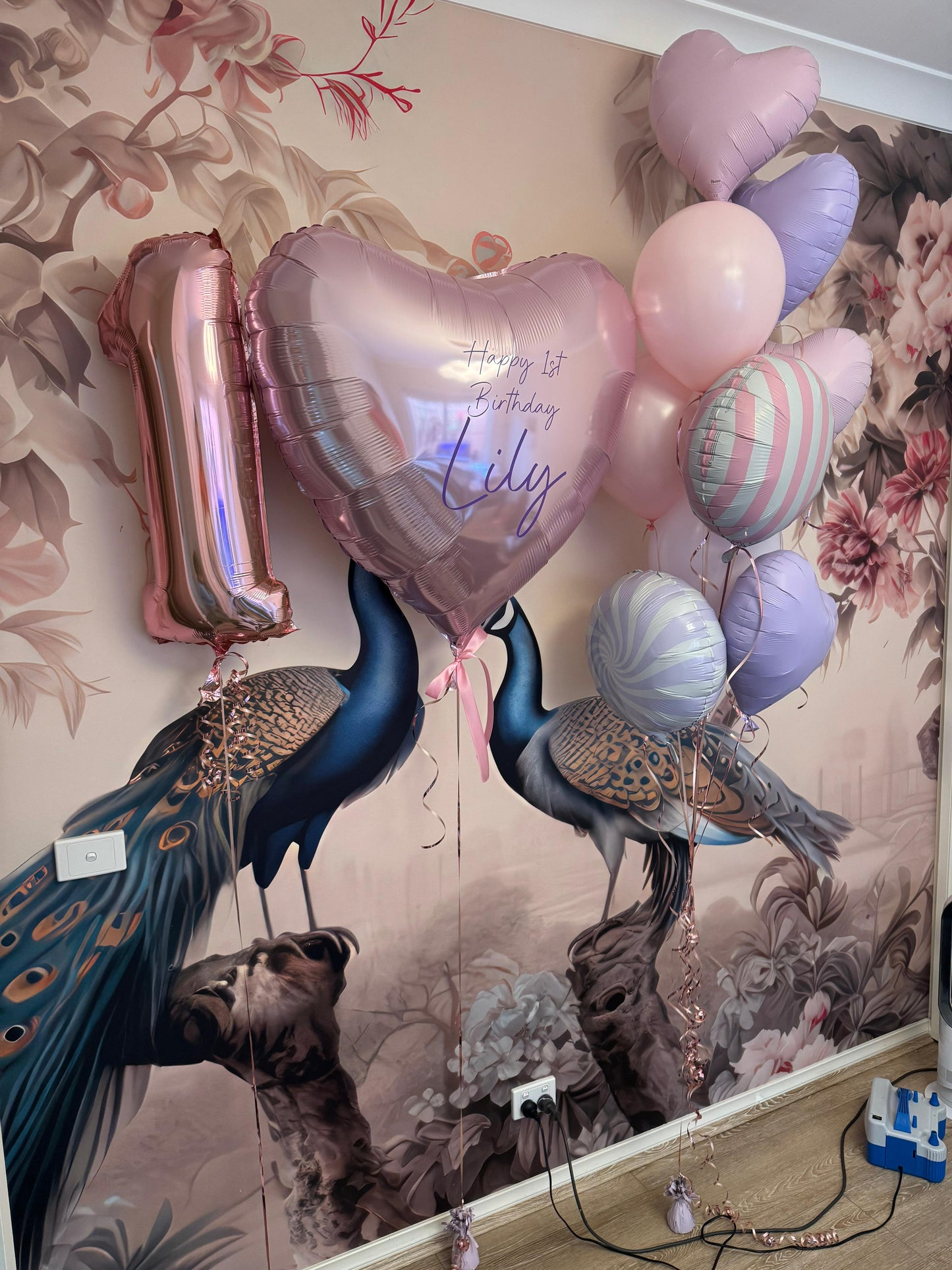Decorative balloons shaped like peacocks and hearts with 'Happy 1st Birthday Lily' text against a floral wall.
