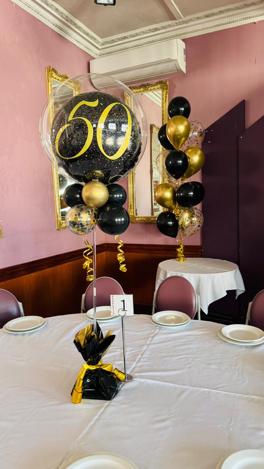 Decorative balloon arrangement with number 50 on a table in a room with pink walls and framed pictures.