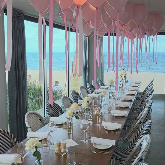 Dining table set for a formal event with pink balloons, white flowers, and striped tablecloths.