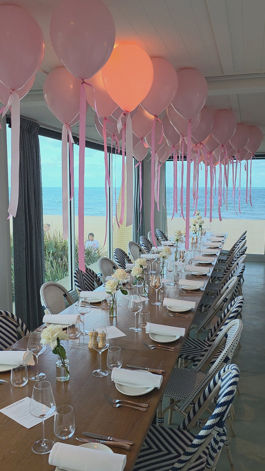 Dining table set for a formal event with pink balloons, white flowers, and striped tablecloths.