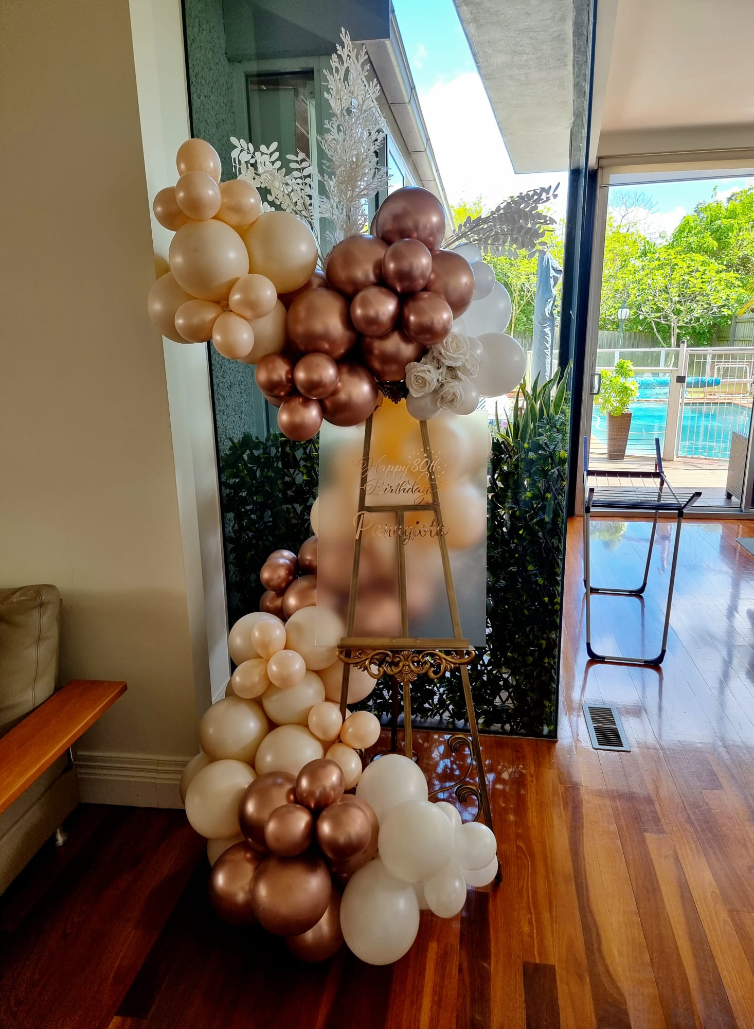 Gold-colored baroque-style easel balloon display with a small balloon garland attached to it, set against an indoor background.