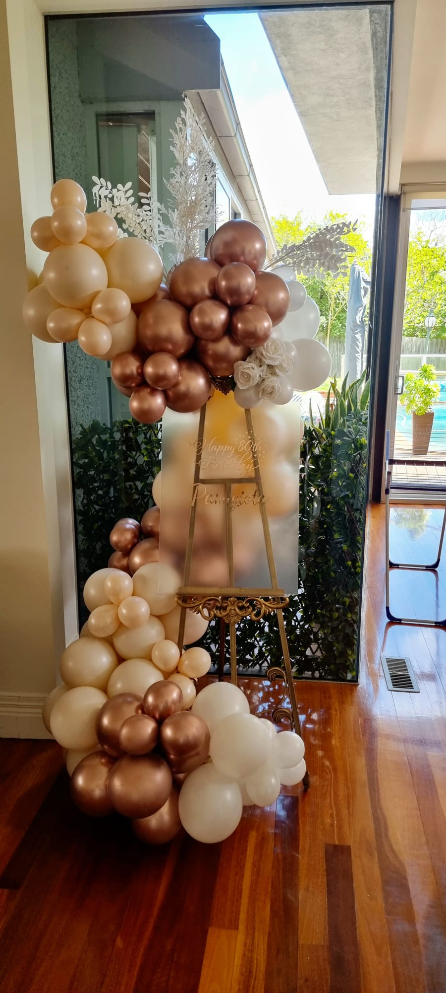 Gold-colored baroque-style easel balloon display with a small balloon garland attached to it, set against an indoor background.